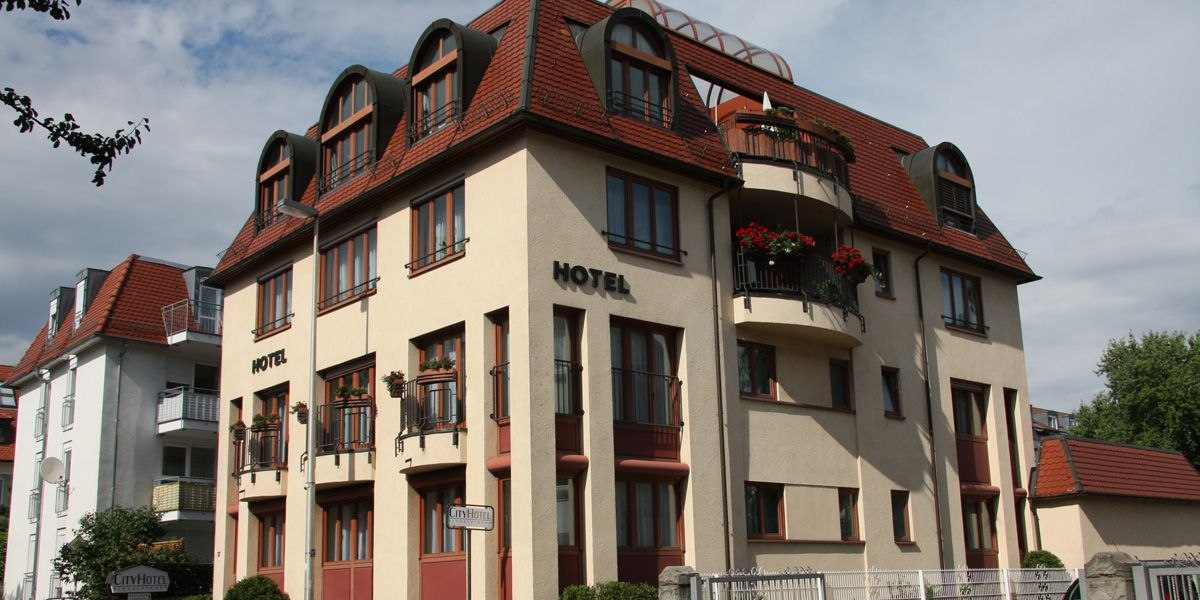 Ein mehrstöckiges Hotel mit roten Dächern und Balkonen, umgeben von anderen Gebäuden und Bäumen., © City Hotel Sindelfingen Ein mehrstöckiges Hotel mit roten Dächern und Balkonen, umgeben von anderen Gebäuden und Bäumen., © City Hotel Sindelfingen