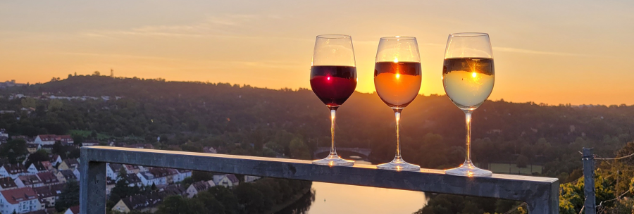 Three wine glasses filled with red, rosé, and white wine are placed on a railing. In the background, the sun is setting over a river landscape., © Weinbau Glock und Sohn GbR
