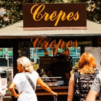 Menschen stehen an einem Crêpes-Stand vor dem Stuttgarter Hauptbahnhof. Der Stand ist mit einem Schild 'Crêpes seit 1980' versehen., © SMG, Sarah Schmid