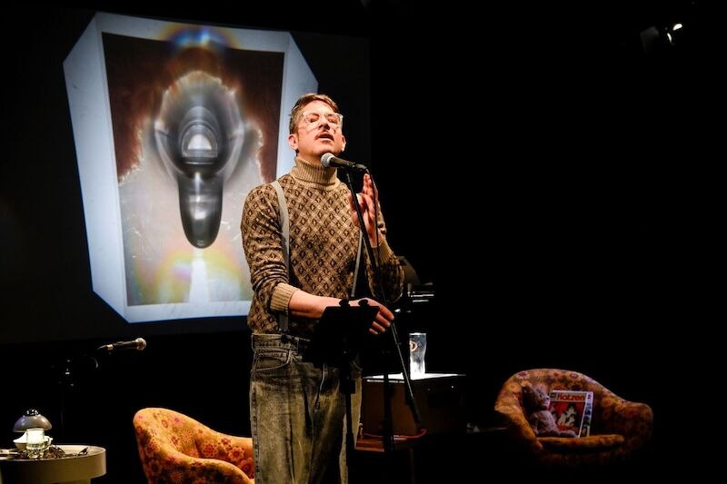 Ein Mann in wei&szlig;em Hemd und Hosentr&auml;gern zeigt auf ein Foto. Im Hintergrund sind bunte Lichter und ein Kleiderst&auml;nder sichtbar., &copy; Theaterhaus Stuttgart e.V.