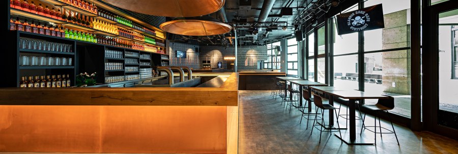 Modernes Café mit Bar, beleuchteten Regalen voller Flaschen, Gläsern und Sitzbereichen. Große Fenster lassen viel Licht herein., © Maks Richter Modernes Café mit Bar, beleuchteten Regalen voller Flaschen, Gläsern und Sitzbereichen. Große Fenster lassen viel Licht herein., © Maks Richter