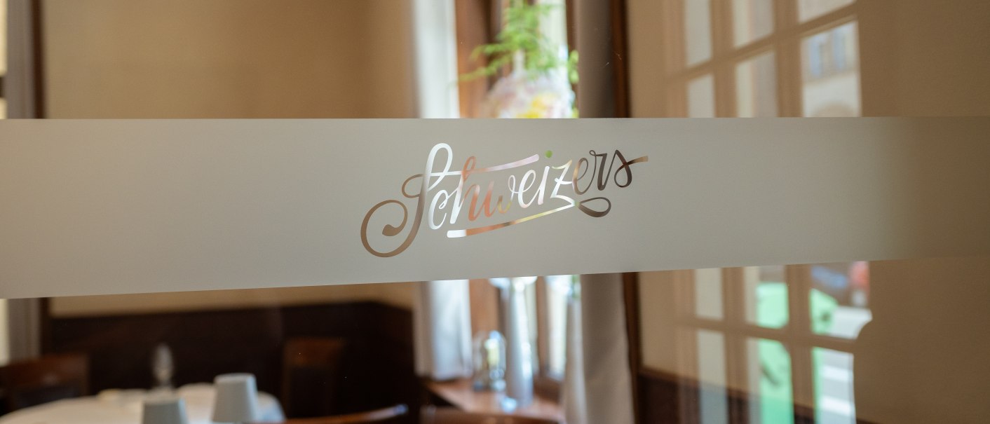 Ein elegantes Restaurant mit dem Schriftzug 'Schweizers' auf einer Glastür, im Hintergrund gedeckte Tische., © Schweizers Restaurant Ein elegantes Restaurant mit dem Schriftzug 'Schweizers' auf einer Glastür, im Hintergrund gedeckte Tische., © Schweizers Restaurant