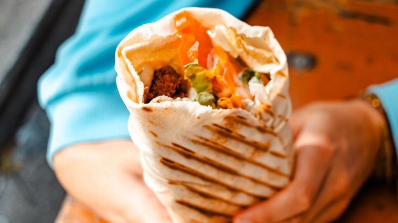 Ein gefüllter Wrap mit Gemüse und Falafel wird von einer Person in blauer Kleidung gehalten., © Stuttgart-Marketing GmbH, Sarah Schmid