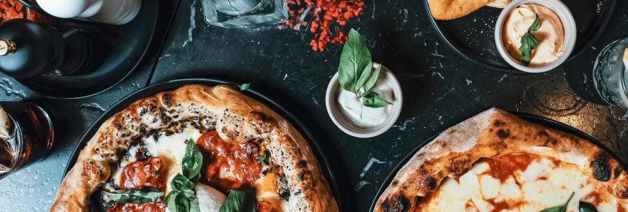 A top-down shot of a table setting featuring several pizzas, dips, glasses, and spice mills on a dark table., &copy; L'Artista, Stuttgart
