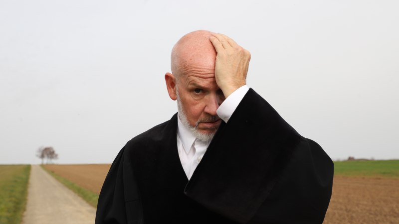 Ein Mann in schwarzer Robe steht auf einem Feldweg und h&auml;lt sich nachdenklich den Kopf. Der Hintergrund zeigt ein leeres Feld und einen Baum., &copy; Sabine Haymann