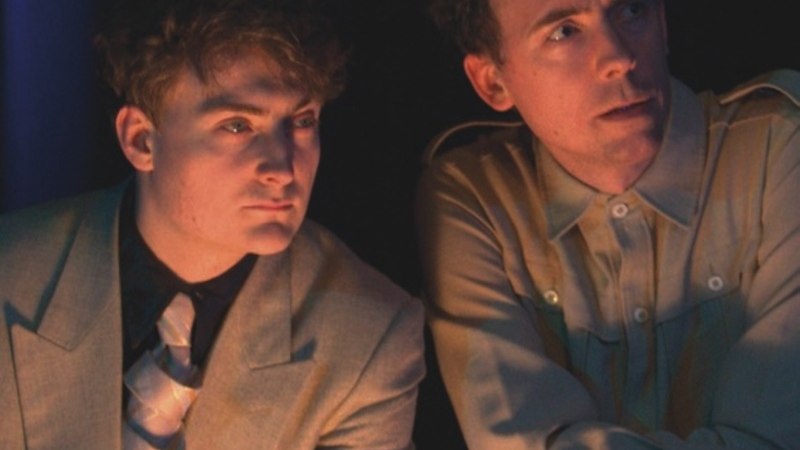 Zwei Männer in Anzügen sitzen nebeneinander und schauen konzentriert in die Ferne. Die Beleuchtung ist gedämpft., © Renitenztheater Stuttgart e.V. Zwei Männer in Anzügen sitzen nebeneinander und schauen konzentriert in die Ferne. Die Beleuchtung ist gedämpft., © Renitenztheater Stuttgart e.V.