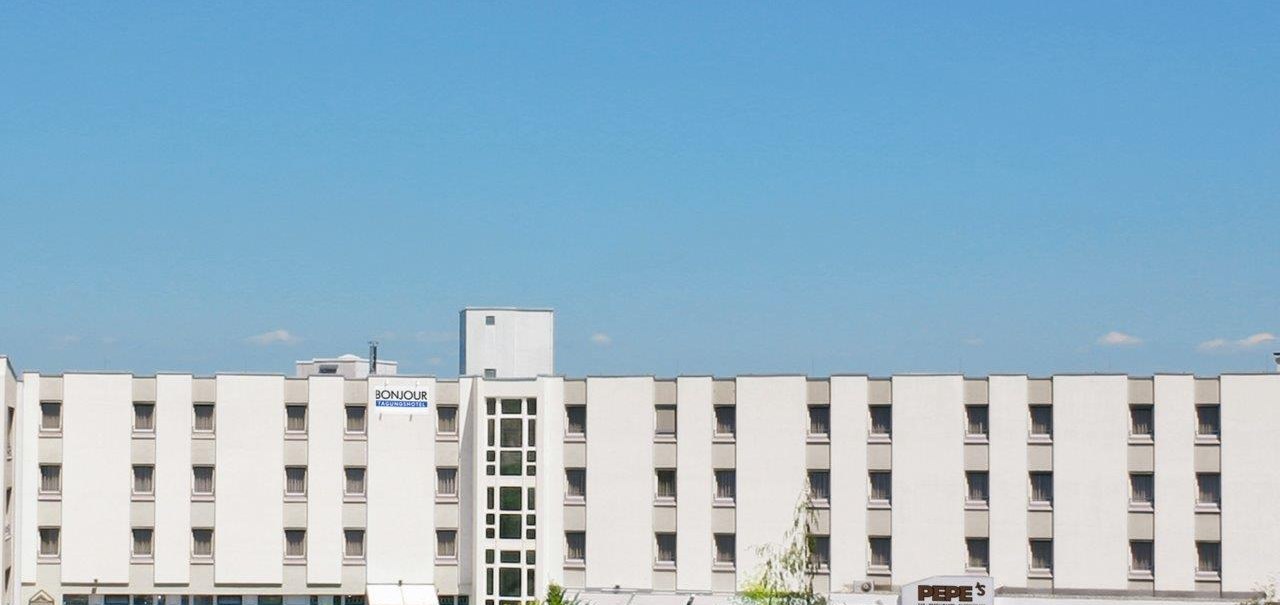 A multi-story hotel building with a white facade and a blue sky in the background. Cars and trees can be seen in front of the hotel., &copy; bonjour Hotelbetriebs GmbH