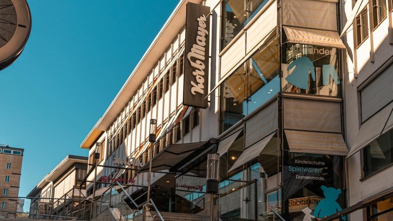 Geb&auml;ude mit Korbmayer-Schild, Schaufenster mit Kinderkleidung und Spielwaren. Moderne Architektur unter blauem Himmel., &copy; Stuttgart Marketing GmbH, Sarah Schmid