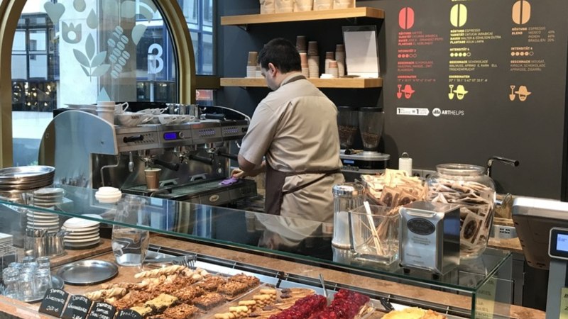 Ein Barista in einem Café bereitet Kaffee zu. Vor ihm liegen Gebäckstücke, daneben eine Kaffeemaschine und Teepackungen., © Old Bridge, Stuttgart