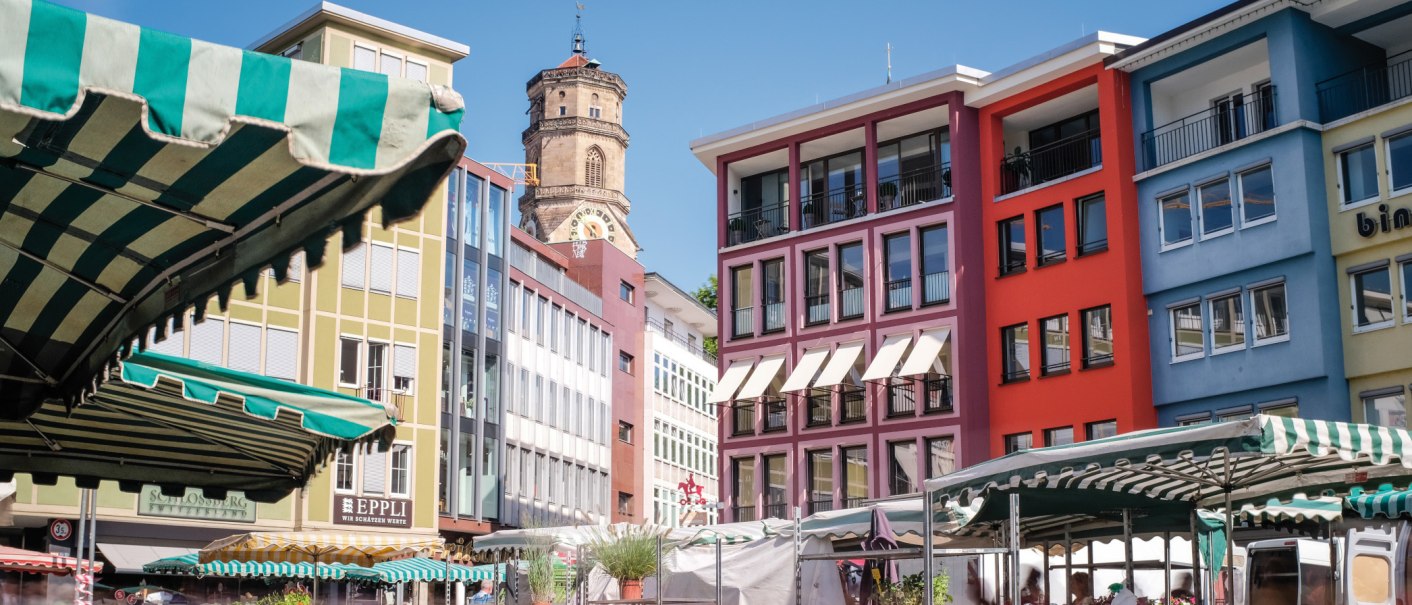 Bunte Gebäude und Marktstände auf einem Marktplatz, mit einem Kirchturm im Hintergrund unter blauem Himmel., © SMG, trickytine Bunte Gebäude und Marktstände auf einem Marktplatz, mit einem Kirchturm im Hintergrund unter blauem Himmel., © SMG, trickytine
