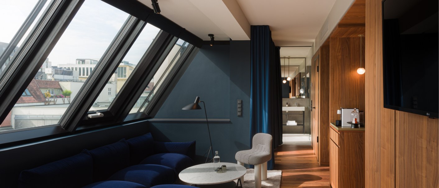 Moderne Suite mit großen Dachfenstern, blauer Couch, Holzboden und Blick ins Badezimmer. Elegantes Design mit Holzelementen und blauen Akzenten., © EmiLu Hotel GmbH