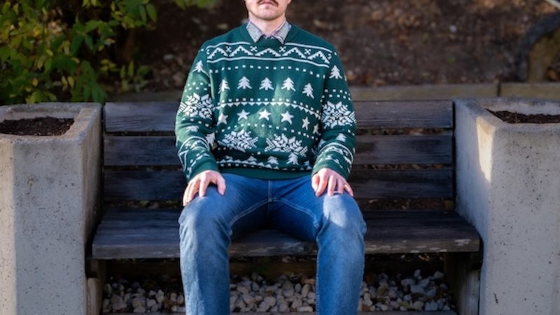 A man with curly hair is sitting on a bench outside. He is wearing a green Christmas sweater with white patterns and blue jeans., © Renitenztheater Stuttgart e.V. A man with curly hair is sitting on a bench outside. He is wearing a green Christmas sweater with white patterns and blue jeans., © Renitenztheater Stuttgart e.V.