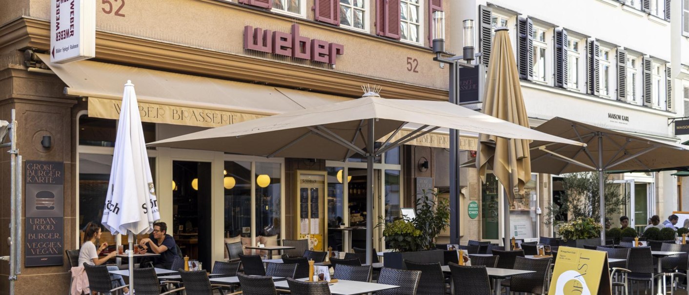 Ein Café mit dem Namen 'Weber' hat einen Außenbereich mit Tischen, Stühlen und Sonnenschirmen. Menschen sitzen und genießen Getränke., © SMG, Sarah Schmid