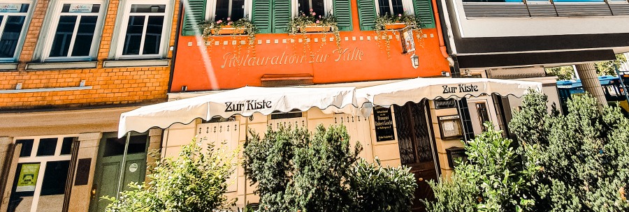 The Zur Kiste wine bar with its red façade, green shutters and awning. Plants adorn the entrance area., © Stuttgart-Marketing GmbH
