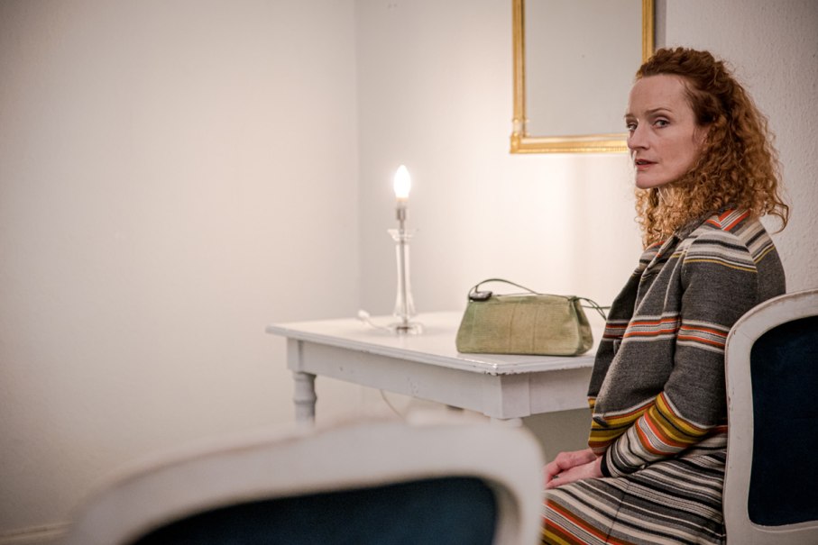 Eine Frau mit lockigem Haar sitzt in einem Raum. Neben ihr ein Tisch mit Lampe und Tasche. Der Raum wirkt schlicht und ruhig., © Thomas Kölsch Eine Frau mit lockigem Haar sitzt in einem Raum. Neben ihr ein Tisch mit Lampe und Tasche. Der Raum wirkt schlicht und ruhig., © Thomas Kölsch