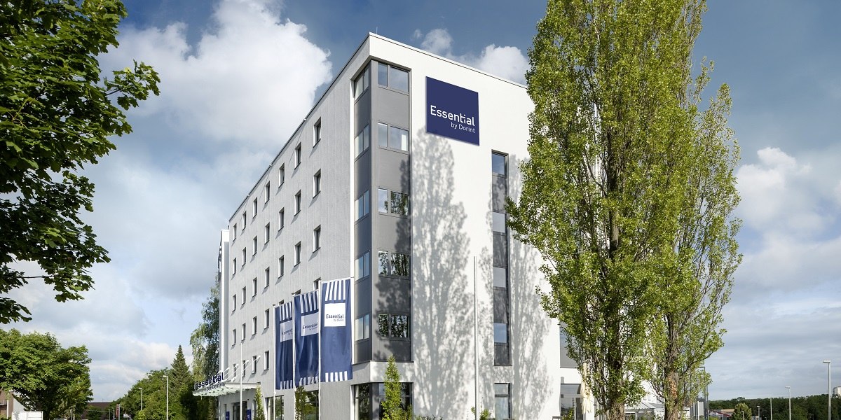 Modernes Hotelgeb&auml;ude mit blauem Logo "Essential by Dorint" und Fahnen, umgeben von B&auml;umen und blauem Himmel., &copy; Essential by Dorint