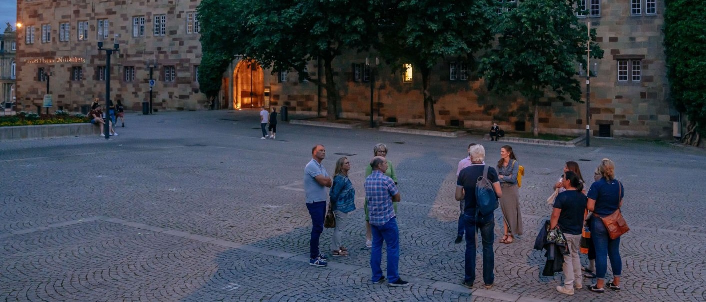Eine Gruppe von Menschen steht auf einem gepflasterten Platz vor einem historischen Gebäude bei Dämmerung., © Thomas Niedermüller