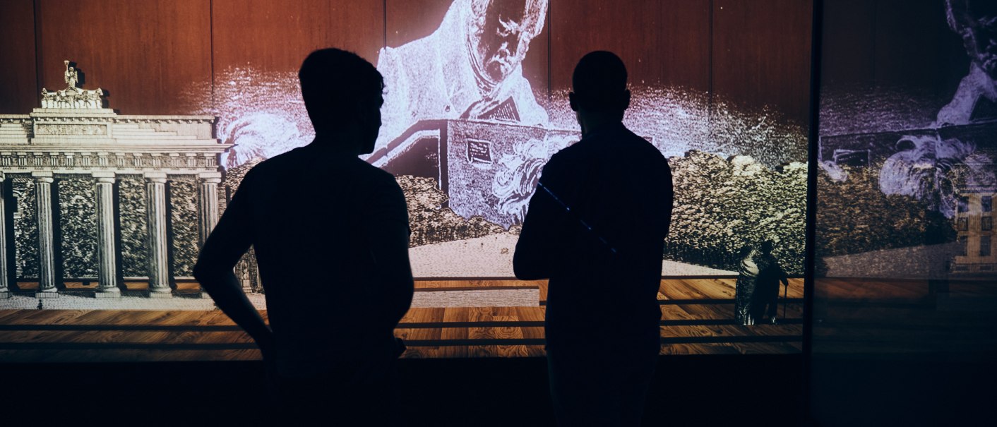 Two people stand in front of a multimedia installation showing Hegel and the Brandenburg Gate. The scene is dark and atmospherically lit., © Julia Ochs