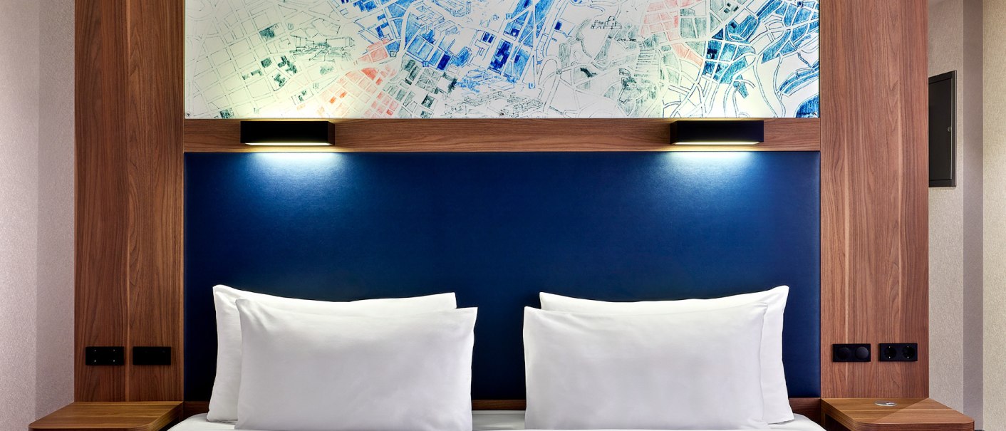 An elegant hotel room with a large bed, blue headboard and stylish wooden accents. Two pillows and a blanket are neatly arranged., © Aloft Stuttgart An elegant hotel room with a large bed, blue headboard and stylish wooden accents. Two pillows and a blanket are neatly arranged., © Aloft Stuttgart