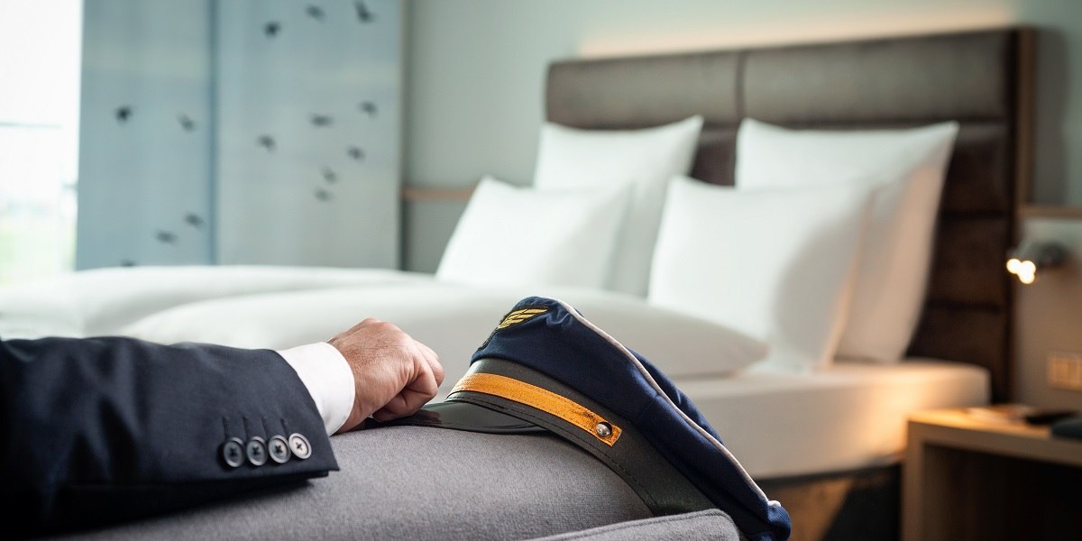 A pilot's jacket and cap lie on a bed in a modern hotel room with white pillows., &copy; Essential by Dorint