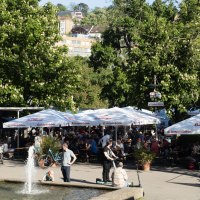 Beer garden in the palace gardens with lots of people under large parasols. A fountain in the foreground, surrounded by green trees and buildings in the background., &copy; Michele Scognamillo