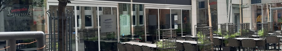 Outdoor area of a restaurant called Enchilada with empty tables and chairs. A tree and plants separate the seating areas., &copy; SMG