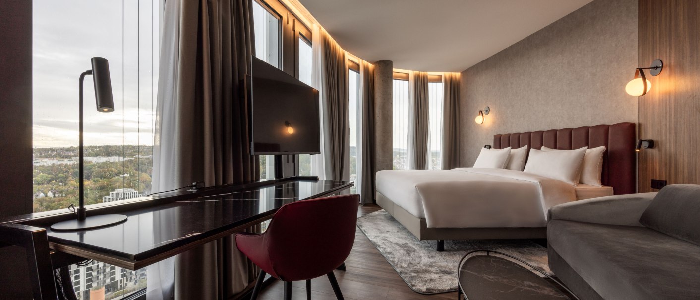 Moderne Hotelsuite mit großem Bett, Schreibtisch, Fernseher und Panoramafenster mit Blick auf die Stadt., © Radisson Hotel Group Moderne Hotelsuite mit großem Bett, Schreibtisch, Fernseher und Panoramafenster mit Blick auf die Stadt., © Radisson Hotel Group