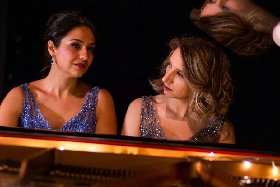 Zwei Frauen in glitzernden Kleidern sitzen nebeneinander an einem Flügel und schauen sich an. Der Hintergrund ist dunkel gehalten., © Blanc&Noir Zwei Frauen in glitzernden Kleidern sitzen nebeneinander an einem Flügel und schauen sich an. Der Hintergrund ist dunkel gehalten., © Blanc&Noir