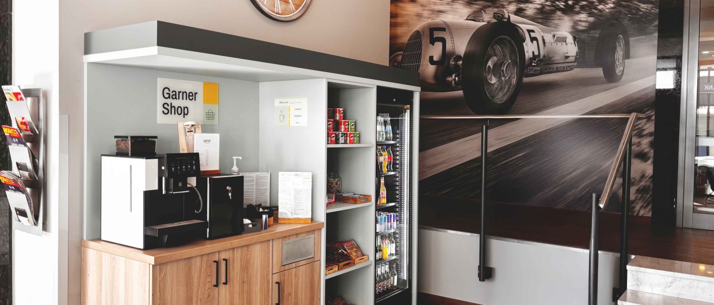 Ein kleiner Shop mit Kaffeemaschine, Snacks und Getr&auml;nken. An der Wand h&auml;ngt ein gro&szlig;es Bild eines Rennwagens. Eine Uhr ist ebenfalls sichtbar., &copy; Novum Hospitality