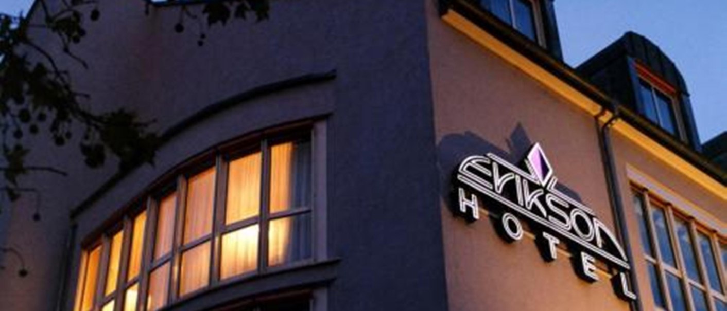 Night view of a hotel with illuminated windows and an illuminated sign with the inscription "Erikson Hotel". Tree branches in the foreground., © Erikson Hotel