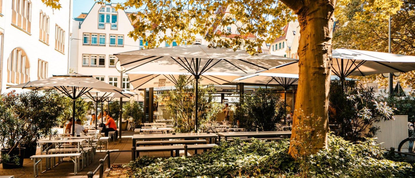 Sonniger Café-Außenbereich mit weißen Sonnenschirmen, Holztischen und Pflanzen. Menschen sitzen entspannt unter den Schirmen. Gebäude im Hintergrund., © Stuttgart Marketing GmbH, Sarah Schmid