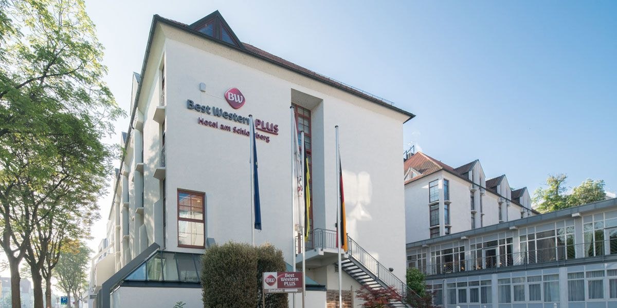 Das Best Western Plus Hotel am Schlossberg mit moderner Fassade, Fahnen und umgeben von Bäumen bei klarem Himmel., © Best Western Plus Hotel am Schlossberg