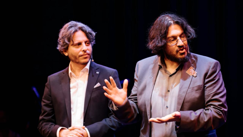 Zwei Männer in Anzügen stehen auf einer Bühne. Der Mann rechts spricht gestikulierend, während der andere aufmerksam zuhört., © Raphael Rodan und Sahand Sahebdivani Zwei Männer in Anzügen stehen auf einer Bühne. Der Mann rechts spricht gestikulierend, während der andere aufmerksam zuhört., © Raphael Rodan und Sahand Sahebdivani