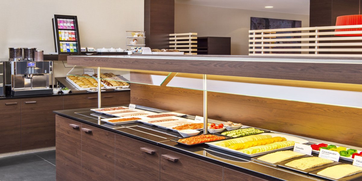 A breakfast buffet featuring a variety of cold cuts, cheeses, pastries, and a coffee machine in a modern, bright room., © NH Ludwigsburg A breakfast buffet featuring a variety of cold cuts, cheeses, pastries, and a coffee machine in a modern, bright room., © NH Ludwigsburg