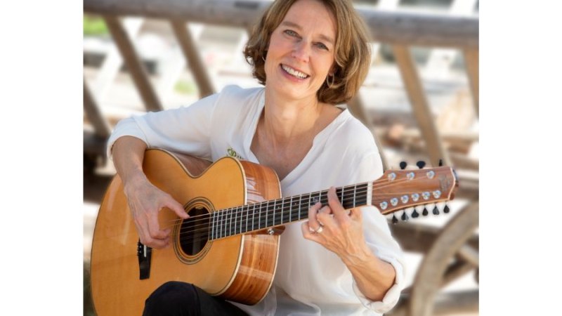 Eine Frau sitzt draußen und spielt Gitarre, während sie in die Kamera lächelt. Sie trägt ein weißes Hemd und wirkt entspannt und glücklich., © Sandra Wolf