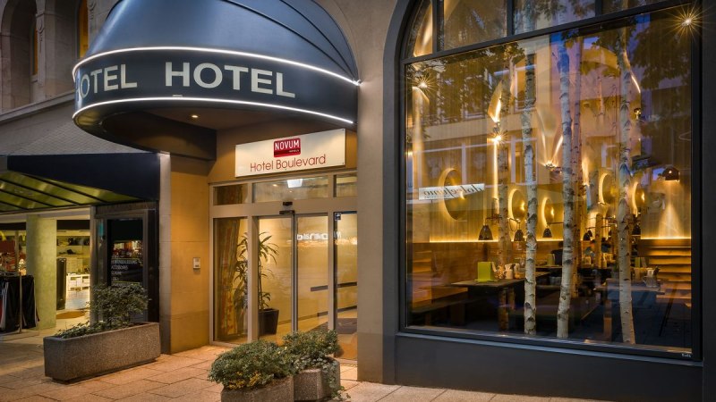 Au&szlig;enansicht eines Hotels mit beleuchtetem Eingang, gro&szlig;em Fenster und Pflanzen. Schild mit der Aufschrift 'Hotel Boulevard' &uuml;ber der T&uuml;r., &copy; Novum Hospitality