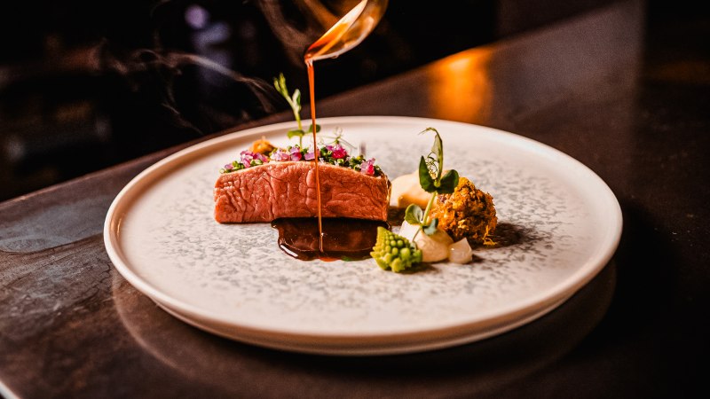 Ein kunstvoll angerichtetes Gourmetgericht mit Fleisch, Gemüse und Sauce auf einem eleganten Teller. Eine Sauce wird darüber gegossen., © wosilatfotografie
