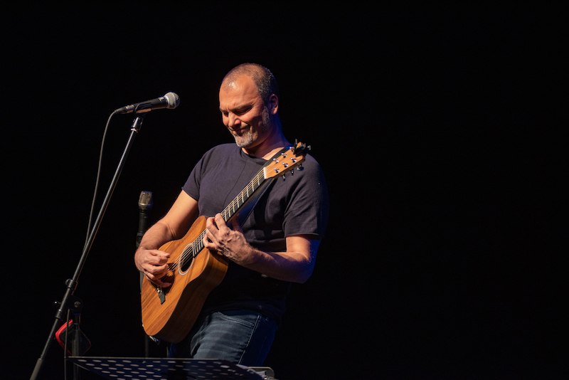 A musician is playing the guitar on a stage, concentrating. He is wearing a dark T-shirt and standing in front of a microphone., © Rosenau Kultur e.V. A musician is playing the guitar on a stage, concentrating. He is wearing a dark T-shirt and standing in front of a microphone., © Rosenau Kultur e.V.