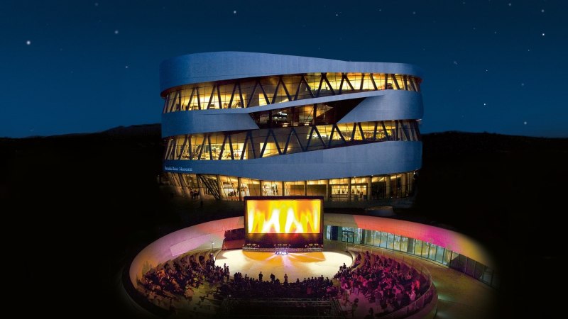 Ein modernes, beleuchtetes Gebäude bei Nacht. Im Vordergrund ein Open-Air-Kino mit Zuschauern und einer großen Leinwand., © Mercedes-Benz Heritage GmbH Ein modernes, beleuchtetes Gebäude bei Nacht. Im Vordergrund ein Open-Air-Kino mit Zuschauern und einer großen Leinwand., © Mercedes-Benz Heritage GmbH