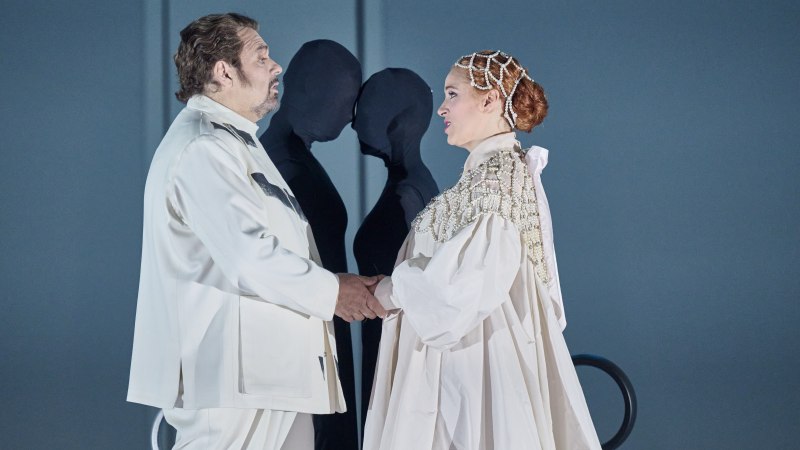 Zwei Opernsänger in weißen Kostümen stehen sich gegenüber und halten sich die Hände. Im Hintergrund sind zwei schwarze Figuren zu sehen., © Martin Sigmund Zwei Opernsänger in weißen Kostümen stehen sich gegenüber und halten sich die Hände. Im Hintergrund sind zwei schwarze Figuren zu sehen., © Martin Sigmund