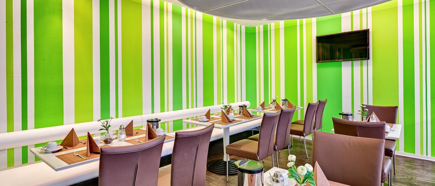 Modern breakfast room with green striped walls, laid tables and brown chairs. A television hangs on the wall., © attimo Hotel Stuttgart Modern breakfast room with green striped walls, laid tables and brown chairs. A television hangs on the wall., © attimo Hotel Stuttgart