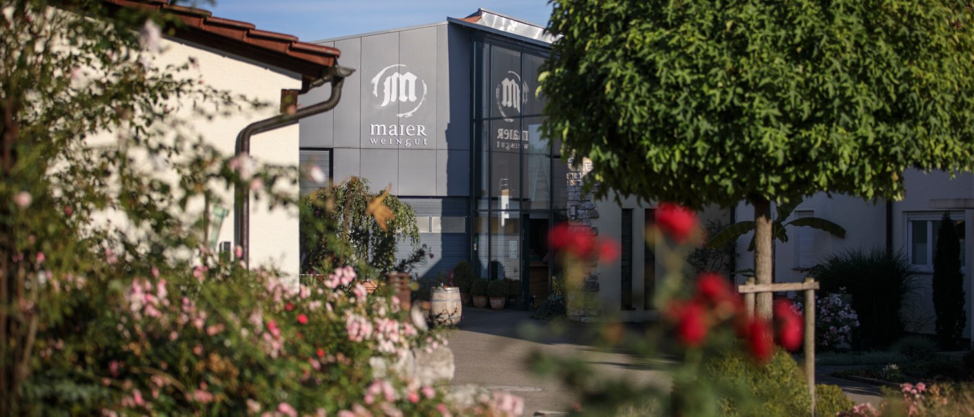 Entrance to the Maier winery, surrounded by flowering plants and trees. Modern architecture with a glass façade and stone walls., © Gottfried Stoppel