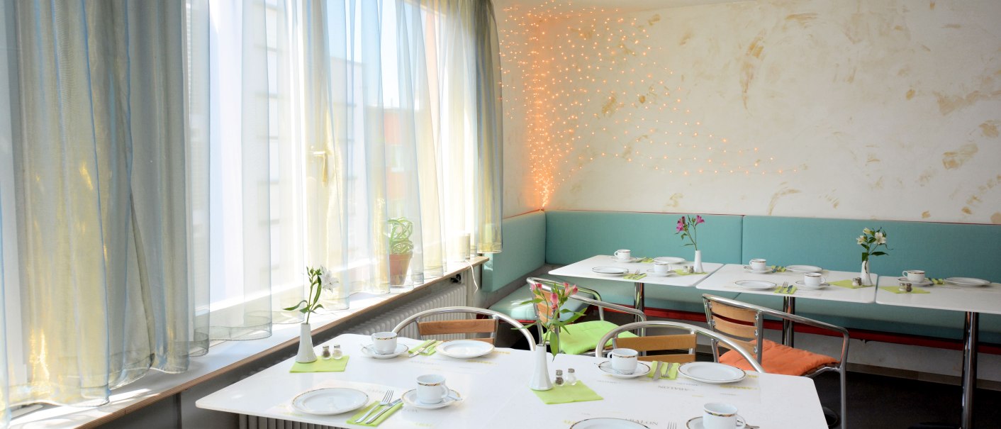 Ein heller Frühstücksraum mit gedeckten Tischen, Blumenvasen und Lichterketten an der Wand. Große Fenster lassen viel Licht herein., © Abalon Hotel Ideal Ein heller Frühstücksraum mit gedeckten Tischen, Blumenvasen und Lichterketten an der Wand. Große Fenster lassen viel Licht herein., © Abalon Hotel Ideal