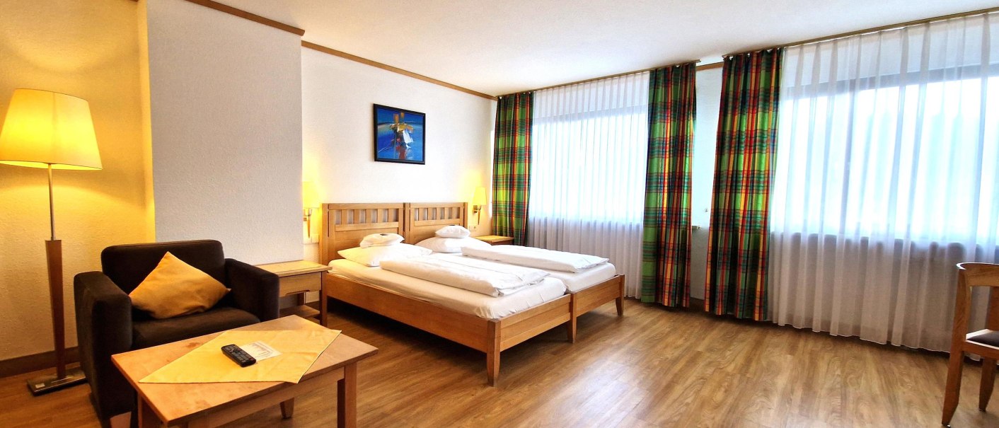 A spacious hotel room with a double bed, an armchair, a table, and large windows with colorful curtains. The hardwood floors and warm lighting create a cozy atmosphere., © TOMAS A spacious hotel room with a double bed, an armchair, a table, and large windows with colorful curtains. The hardwood floors and warm lighting create a cozy atmosphere., © TOMAS