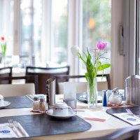 Ein Tisch in einem Caf&eacute; ist mit Geschirr, Besteck und einer Vase mit Tulpen gedeckt. Im Hintergrund sind Fenster und weitere Tische zu sehen., &copy; TOMAS