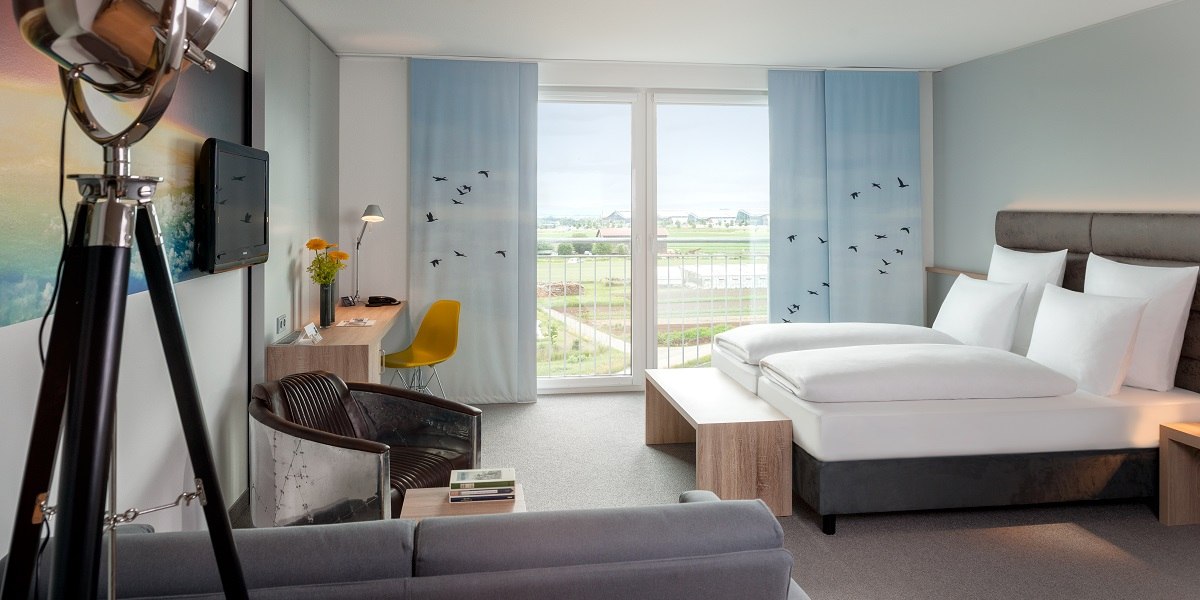 Modern hotel room with large bed, desk, armchair and balcony with a view of the countryside. Decor with blue curtains and bird motifs., &copy; Essential by Dorint