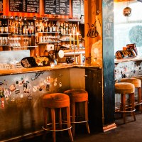Gemütliche Bar mit Barhockern, einer Theke voller Flaschen und Gläser. Die Wände sind mit Stickern dekoriert, und eine Tafel zeigt Getränkepreise., © Mata Hari Gemütliche Bar mit Barhockern, einer Theke voller Flaschen und Gläser. Die Wände sind mit Stickern dekoriert, und eine Tafel zeigt Getränkepreise., © Mata Hari