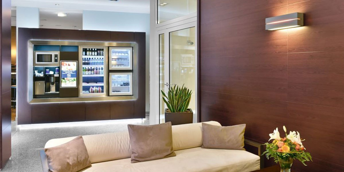 Moderne Lobby mit beigem Sofa, Pflanzen und beleuchtetem Snackautomaten. Holzwände und dezente Beleuchtung schaffen eine einladende Atmosphäre., © NH Stuttgart Sindelfingen Moderne Lobby mit beigem Sofa, Pflanzen und beleuchtetem Snackautomaten. Holzwände und dezente Beleuchtung schaffen eine einladende Atmosphäre., © NH Stuttgart Sindelfingen
