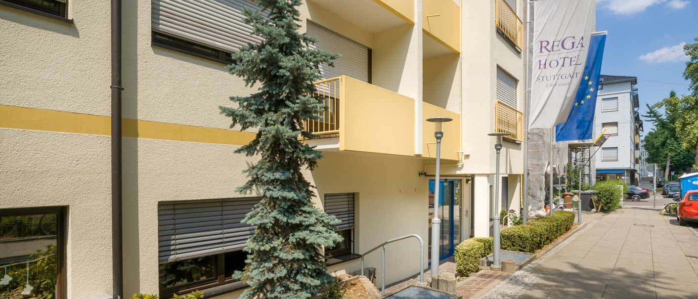 Exterior view of a hotel with yellow balconies, a blue door and flags. A tree and plants line the path along the building., © TOMAS