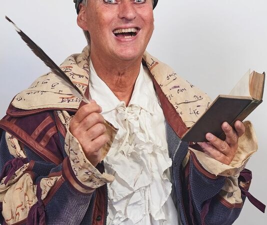 Person in bunter, historischer Kleidung mit Krone, hält eine Feder und ein Buch, lächelt breit in die Kamera., © Theaterhaus Stuttgart e.V.